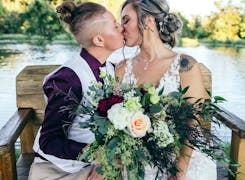 A freshly married couple, wedding bouquet between them, share a kiss beside the water