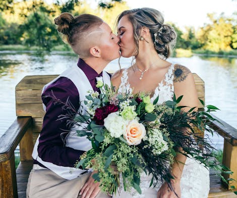 A freshly married couple, wedding bouquet between them, share a kiss beside the water
